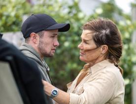 A production still from the series After the Party filmed in Wellington. Penny (played by Robyn Malcolm) wears a beige blouse and stands facing Tom (played by Elz Carrad) who wears a black baseball cap with a distressed facial expression.