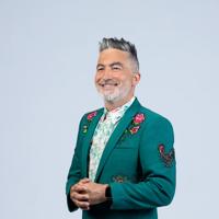 Kiwi comedian, Dai Henwood, looks sharp wearing a dark green suit jacket with embroidered roses and a rooster on a light grey backdrop.