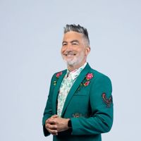 Kiwi comedian, Dai Henwood, looks sharp wearing a dark green suit jacket with embroidered roses and a rooster on a light grey backdrop.