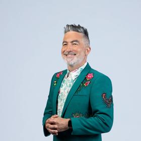 Kiwi comedian, Dai Henwood, looks sharp wearing a dark green suit jacket with embroidered roses and a rooster on a light grey backdrop.