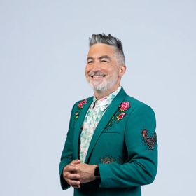 Kiwi comedian, Dai Henwood, looks sharp wearing a dark green suit jacket with embroidered roses and a rooster on a light grey backdrop.
