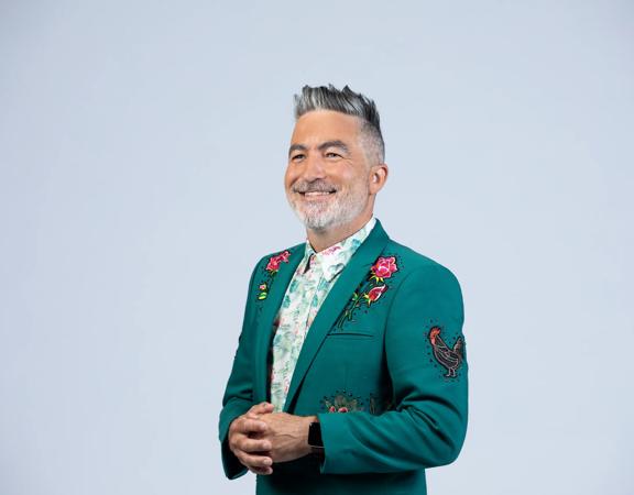 Kiwi comedian, Dai Henwood, looks sharp wearing a dark green suit jacket with embroidered roses and a rooster on a light grey backdrop.
