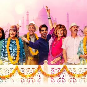 Seven actors in the stage production of The Best Exotic Marigold Hotel wear summer holiday outfits while standing on a balcony. 