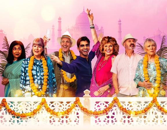 Seven actors in the stage production of The Best Exotic Marigold Hotel wear summer holiday outfits while standing on a balcony. 
