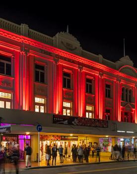 The exterior of Wellington's The Opera House at night. The building's facade is lit up in red and people assemble outside the venue.