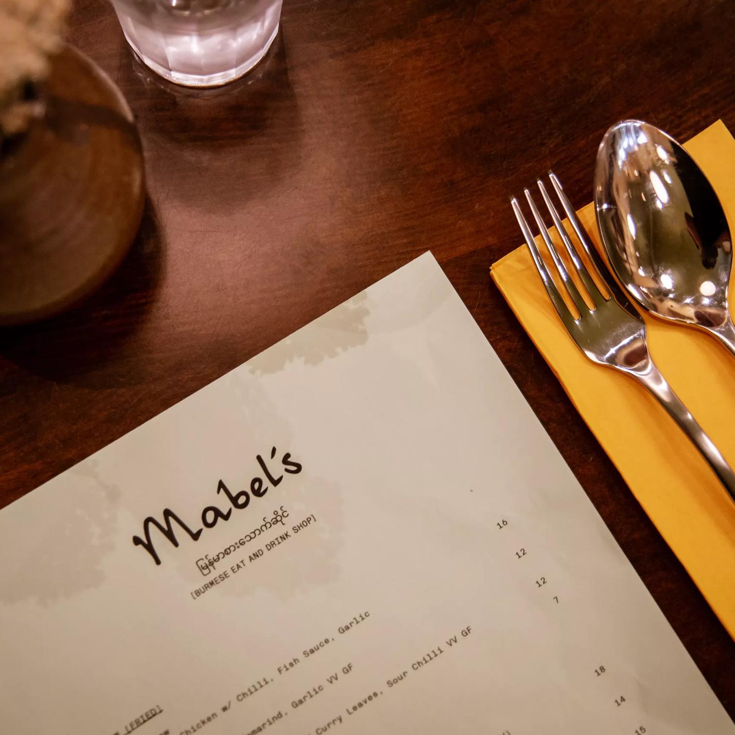 The menu at Mabel's, a Burmese Eat & Drink Shop in Wellington's city centre, placed on a wooden table with a brown flower vase, a water glass, and a yellow napkin with a fork and spoon. 