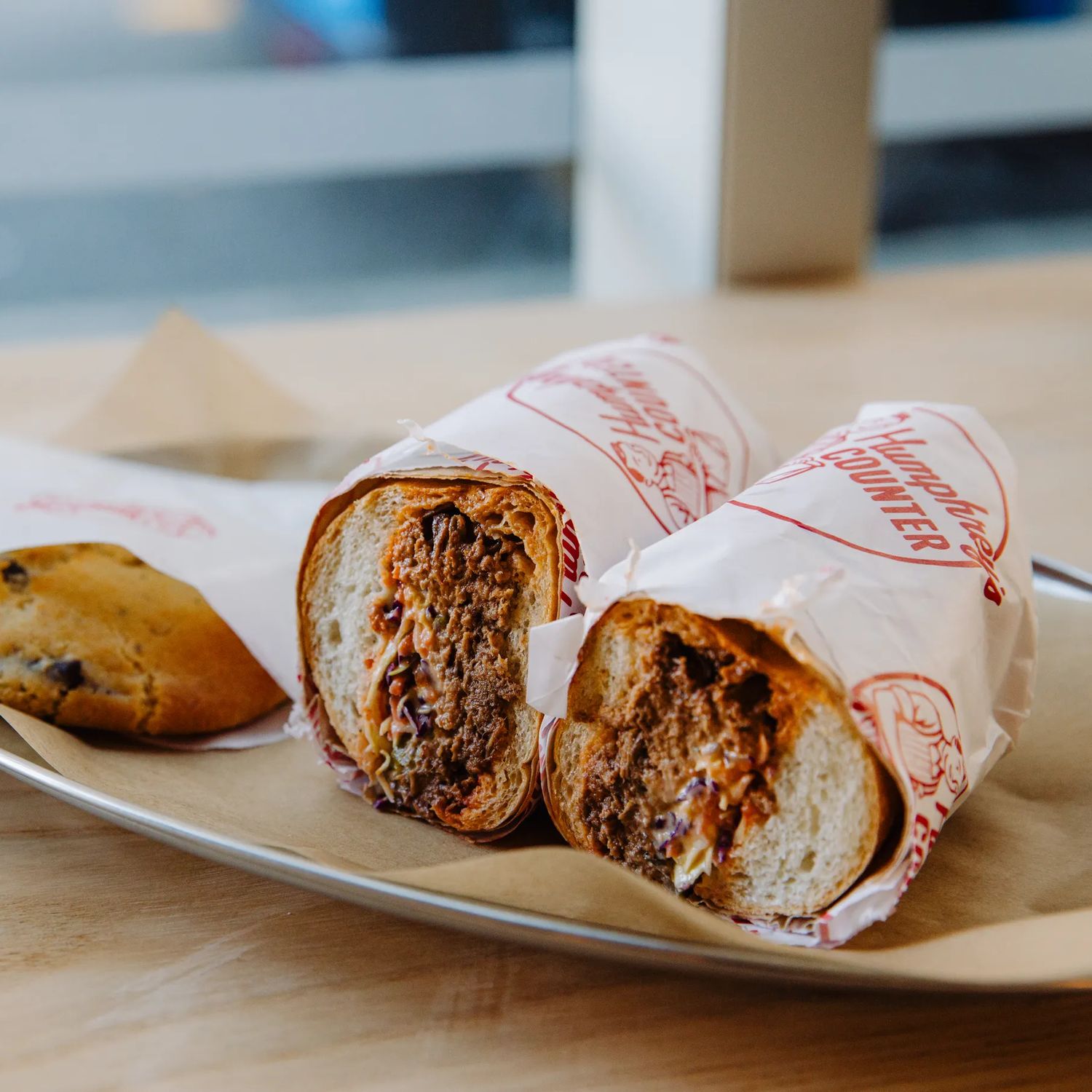 A toasted filled wrap cut in half and served on a metal tray beside a small pastry, on a light wooden table with a softly blurred café background.