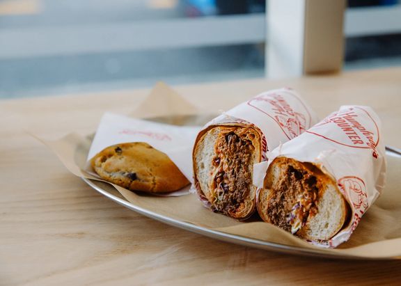 A toasted filled wrap cut in half and served on a metal tray beside a small pastry, on a light wooden table with a softly blurred café background.