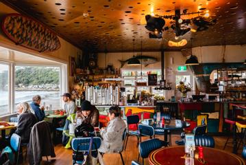 Customers sitting inside Maranui Cafe enjoying the lookout over Lyall Bay and their meals. Fish and surf decor surround the room.