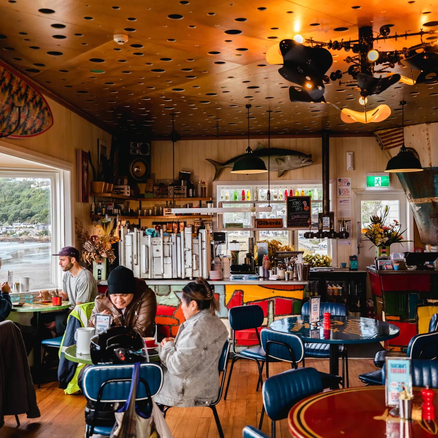 Customers sitting inside Maranui Cafe enjoying the lookout over Lyall Bay and their meals. Fish and surf decor surround the room.