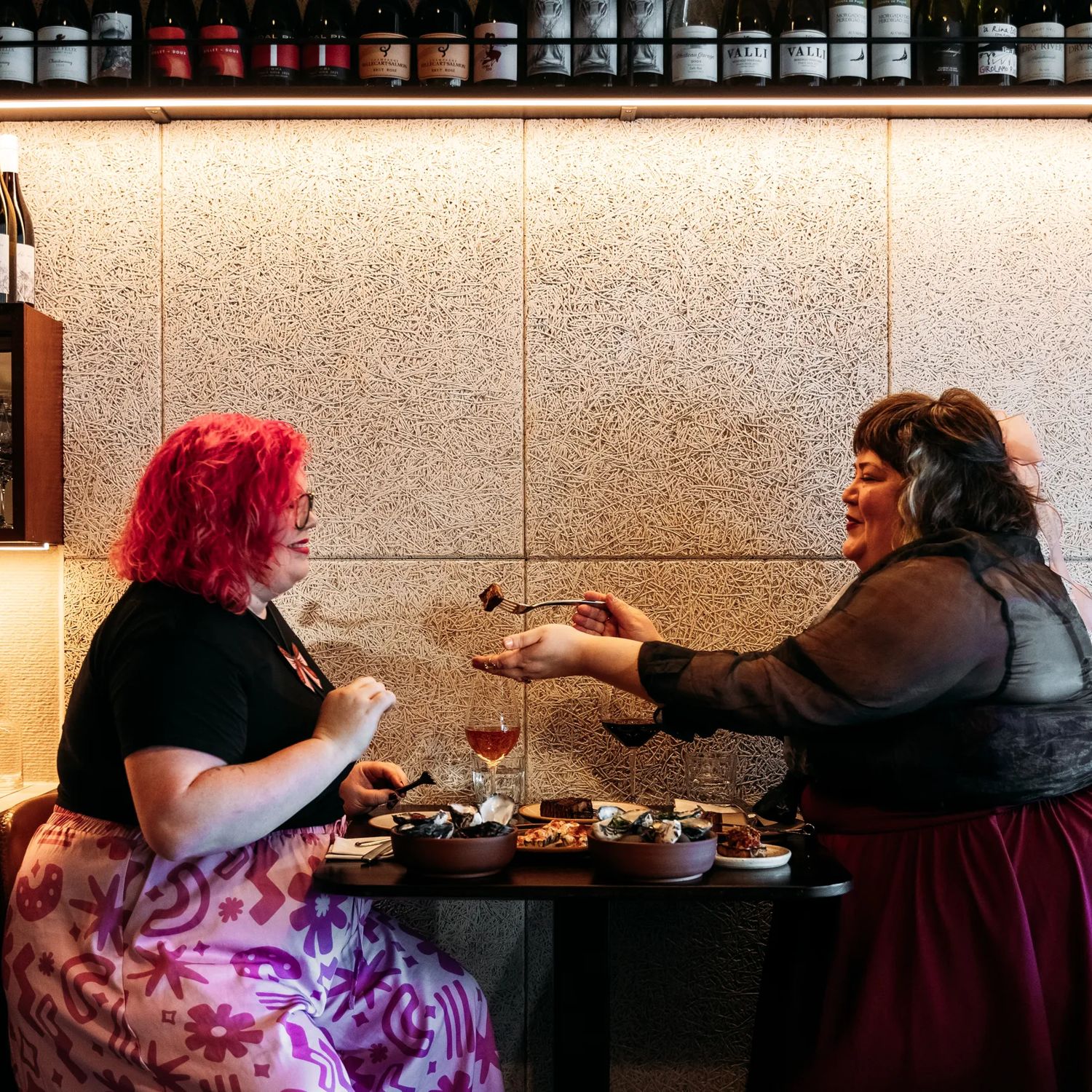 Two people at Rosella eating food. A rack of wine bottles is above them.