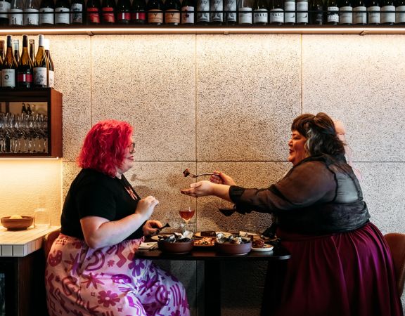 Two people at Rosella eating food. A rack of wine bottles is above them.