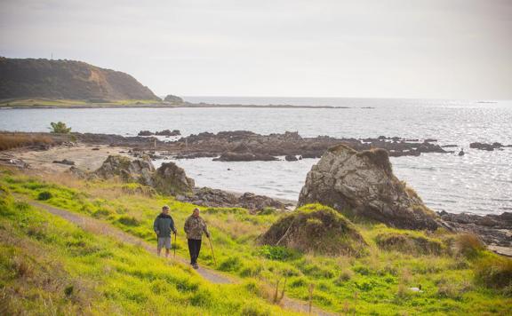 Two people using walking sticks are strolling along a gravel path next to a rocky shoreline.