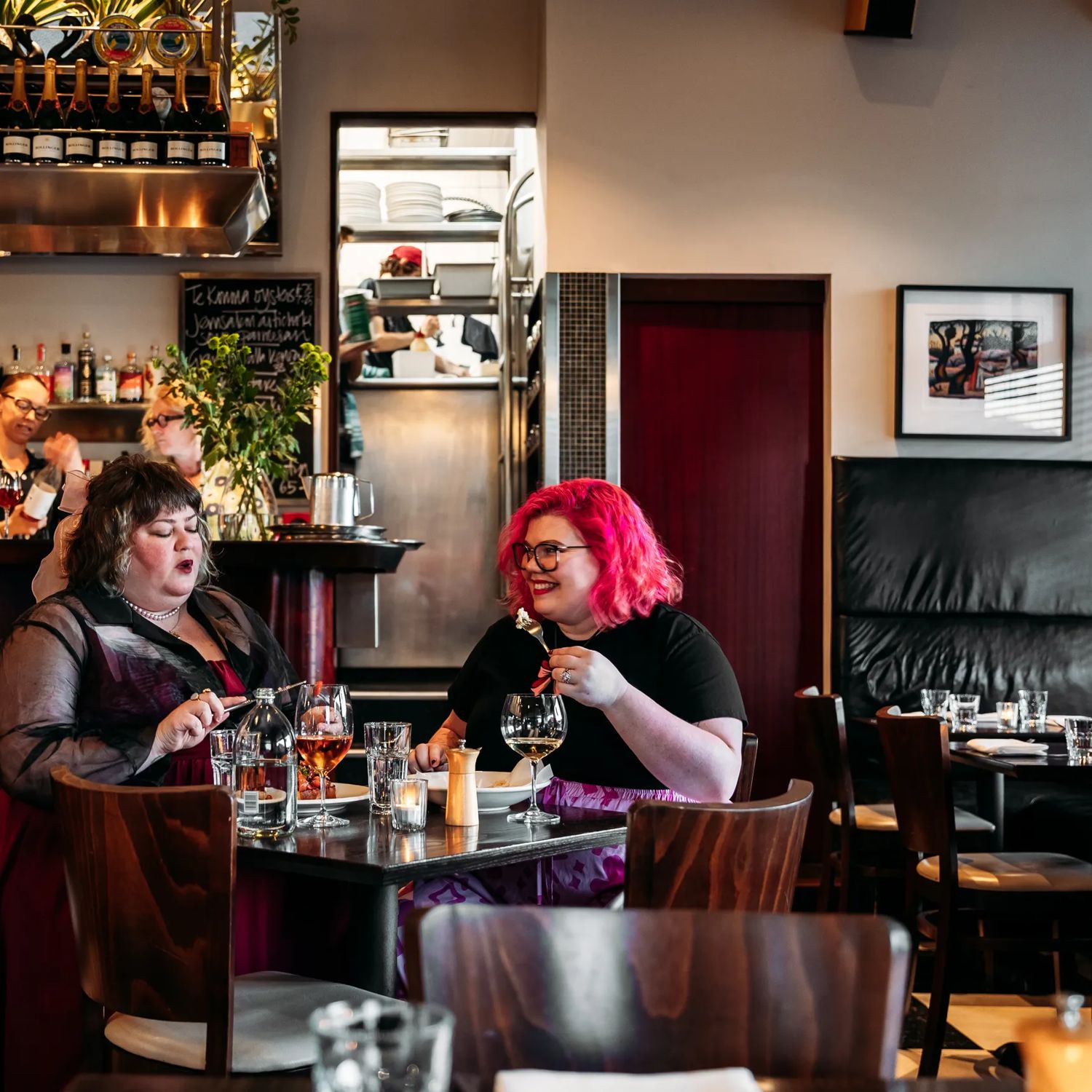 Two people enjoying a meal in Capitol Restaurant while staff make drinks behind the bar behind them. The dark wood tables add a moody vibe to the interior.