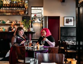 Two people enjoying a meal in Capitol Restaurant while staff make drinks behind the bar behind them. The dark wood tables add a moody vibe to the interior.