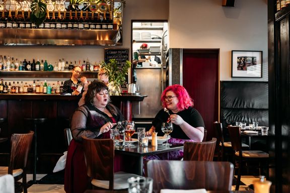Two people enjoying a meal in Capitol Restaurant while staff make drinks behind the bar behind them. The dark wood tables add a moody vibe to the interior.