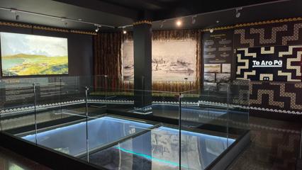 Inside an exhibition space in Te Wharewaka o Pōneke, a multipurpose function centre and waka house located on Taranaki Stree Wharf on Wellington's waterfront. The dark room has images and text on display with a glass floor in the middle of the space.