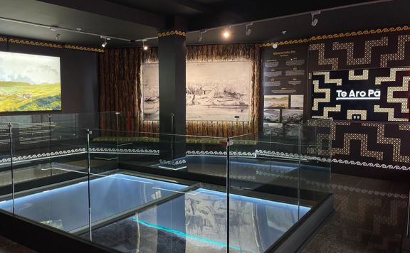 Inside an exhibition space in Te Wharewaka o Pōneke, a multipurpose function centre and waka house located on Taranaki Stree Wharf on Wellington's waterfront. The dark room has images and text on display with a glass floor in the middle of the space.