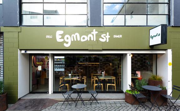 The exterior of Egmont St Deli & Diner. The Brick wall is painted green with a couple of small tables and chairs set up outside. 