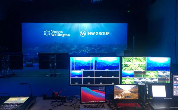 A control room for Venues Wellington Virtual Events. THe room is dark with several computer monitors, laptops, a projection screen and other technical equipment.