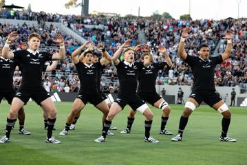 The New Zealand All Blacks rugby team perform their haka on the field with sports fans watching from the stands.