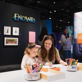 A smiling adult sits with a child drawing with coloured pencils at a table.