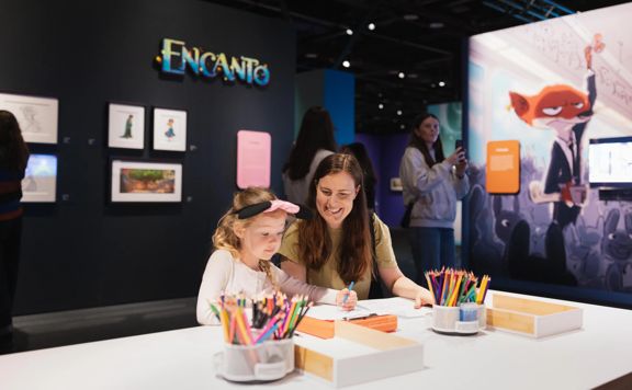 A smiling adult sits with a child drawing with coloured pencils at a table.