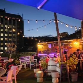 The Arborist's rooftop terrace in the evening with people sitting, eating and drinking with string lights above and buildings in the background.