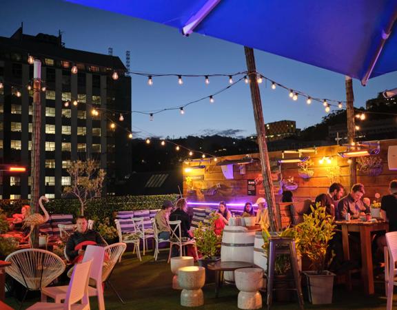 The Arborist's rooftop terrace in the evening with people sitting, eating and drinking with string lights above and buildings in the background. 