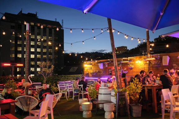 The Arborist's rooftop terrace in the evening with people sitting, eating and drinking with string lights above and buildings in the background.