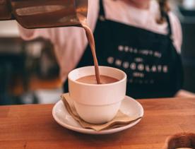 A person wearing a white T-shirt and a black apron is pouring a hot chocolate into a small white mug at the Wellington Chocolate Factory. 