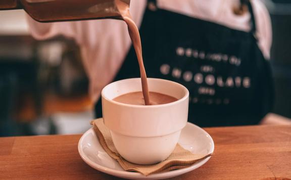 A person wearing a white T-shirt and a black apron is pouring a hot chocolate into a small white mug at the Wellington Chocolate Factory.